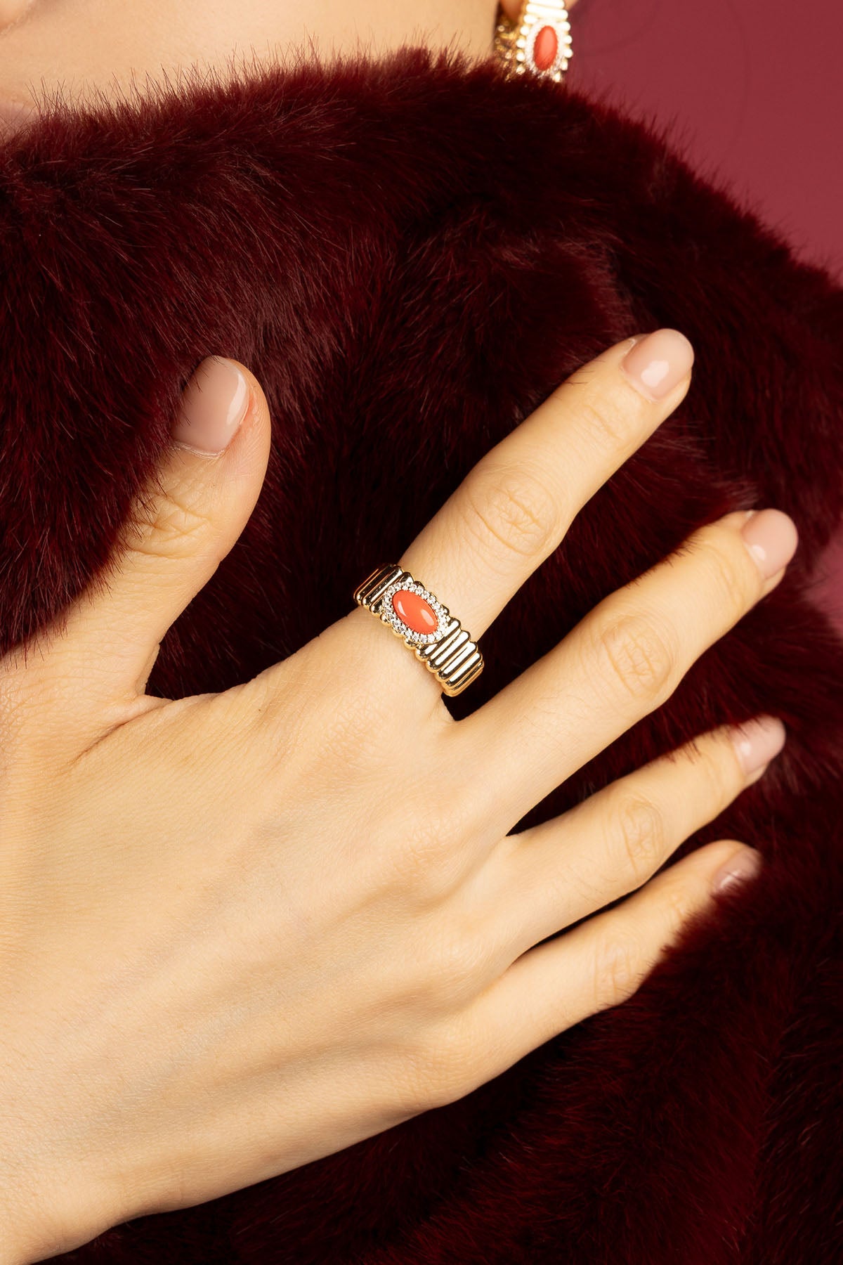 Woman wearing APM Monaco ring, coral warmth against burgundy fur, quiet winter elegance.
