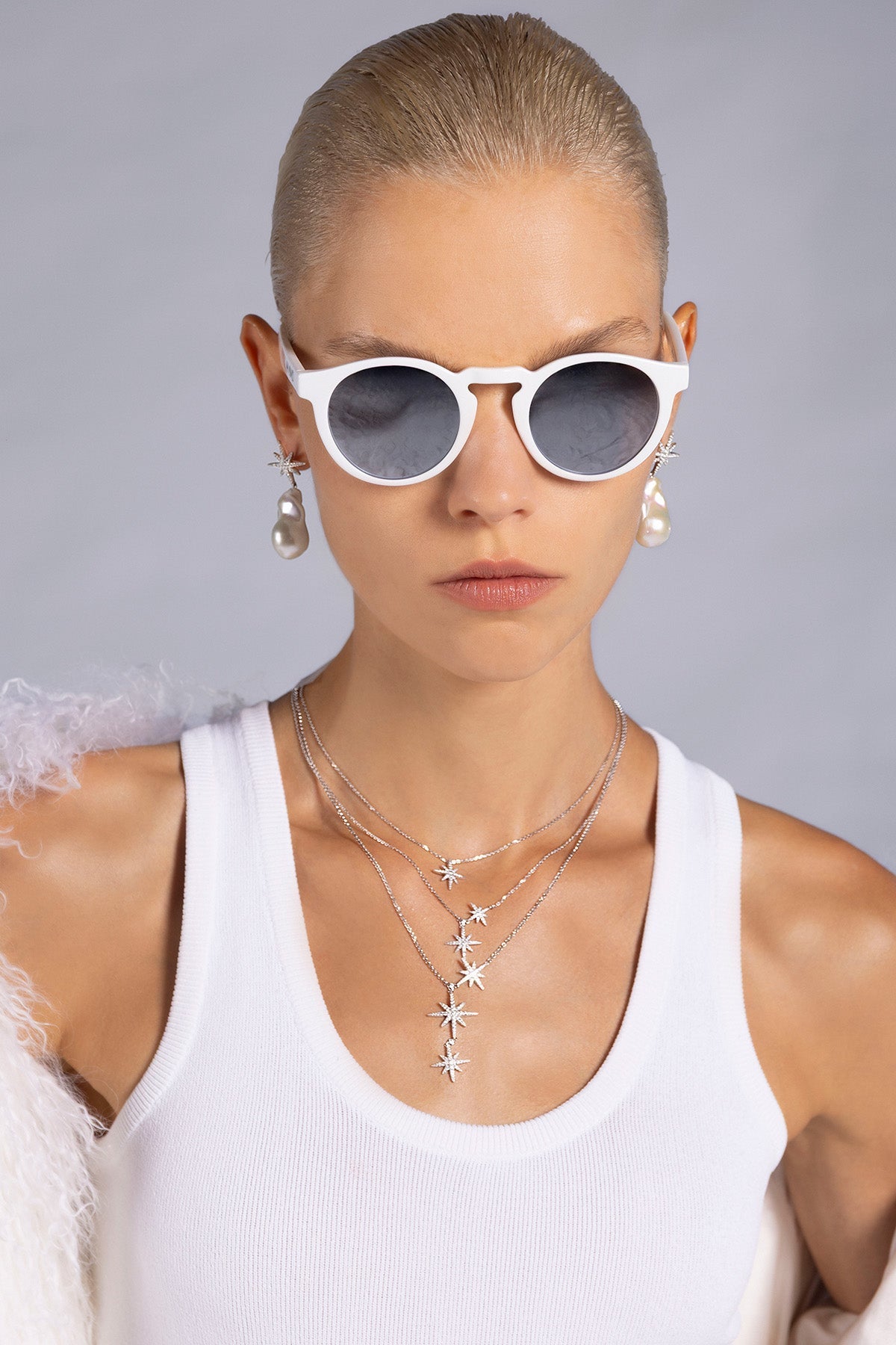 Woman wearing white sunglasses and layered necklaces against a gray background