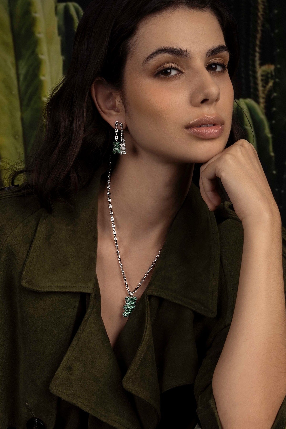 Woman wearing a green jacket with jewelry, including earrings and a necklace, against a blurred natural background.