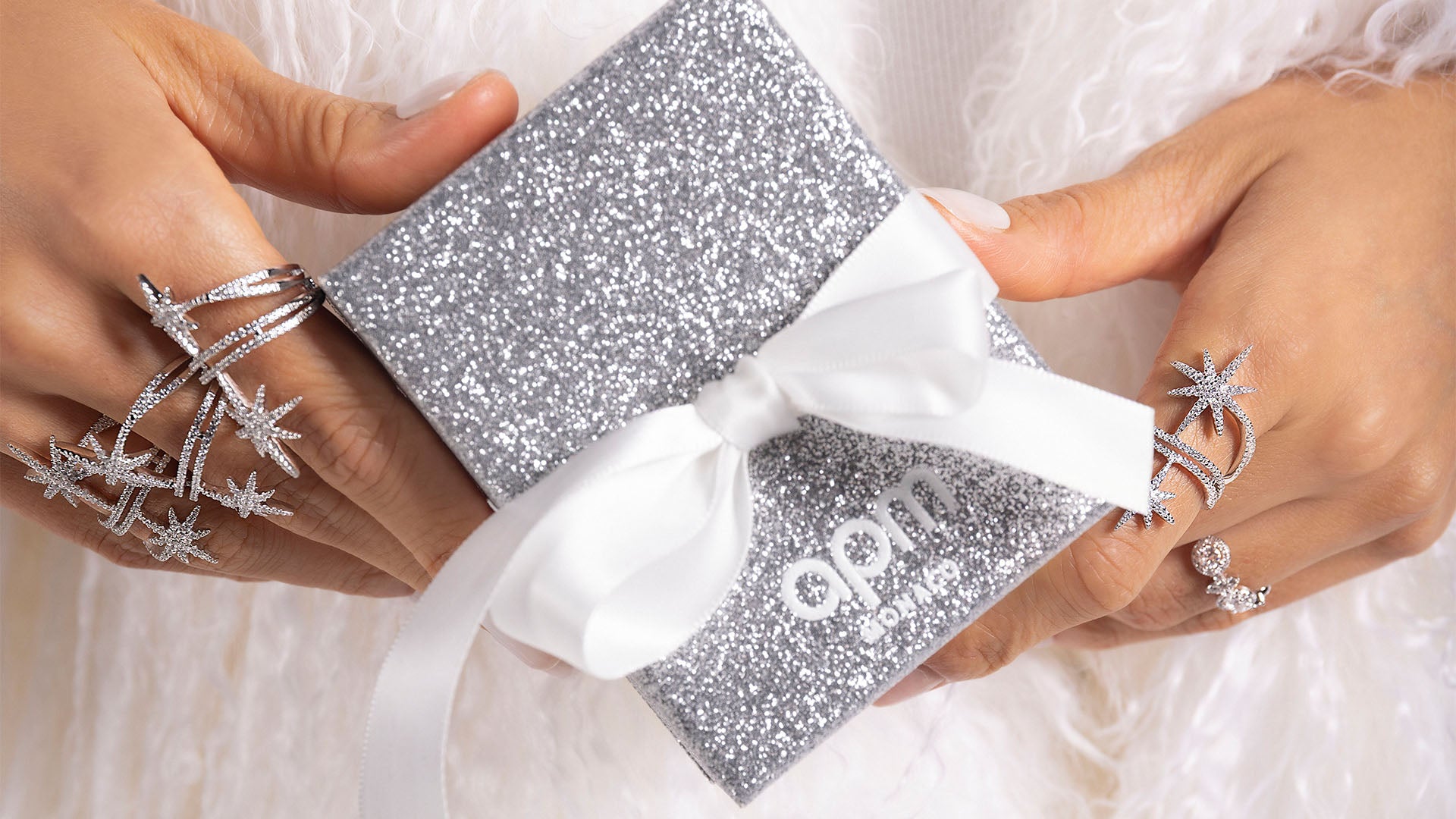 Silver glittery gift box with white ribbon held by hands, featuring the brand 'APM MONACO'.