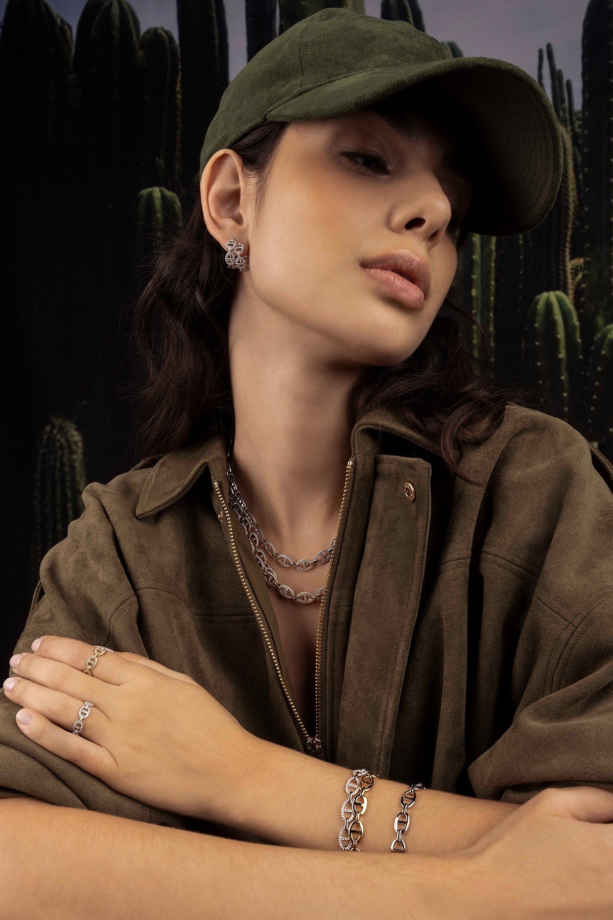 Woman wearing a green cap and brown jacket with jewelry against a cactus background