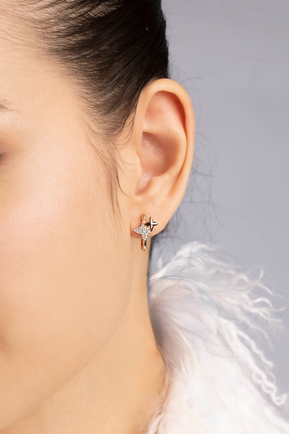 Close-up of an ear wearing a rose gold earring with a clear gemstone against a neutral background