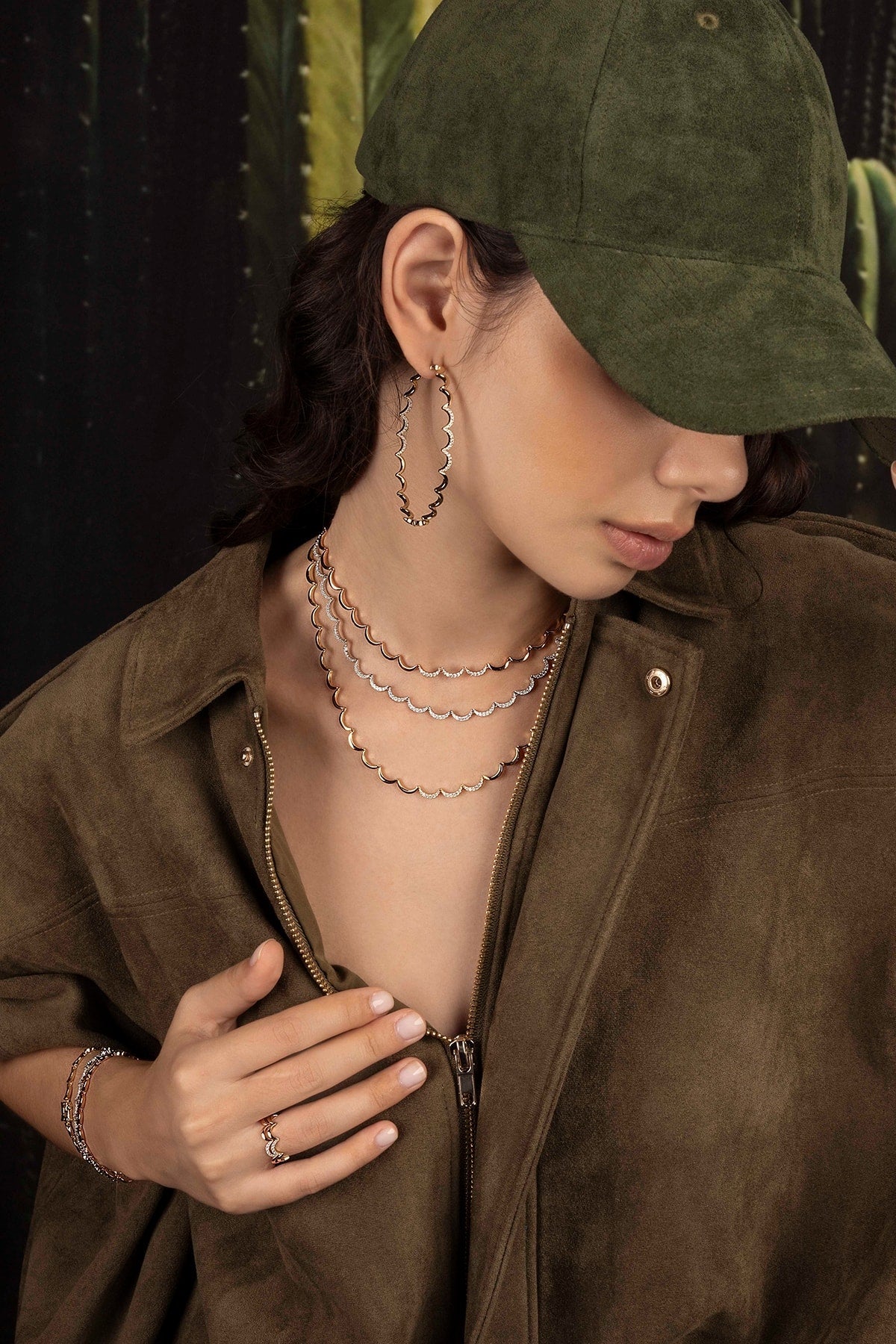 Model wearing a green cap and brown jacket with layered necklaces against a dark background