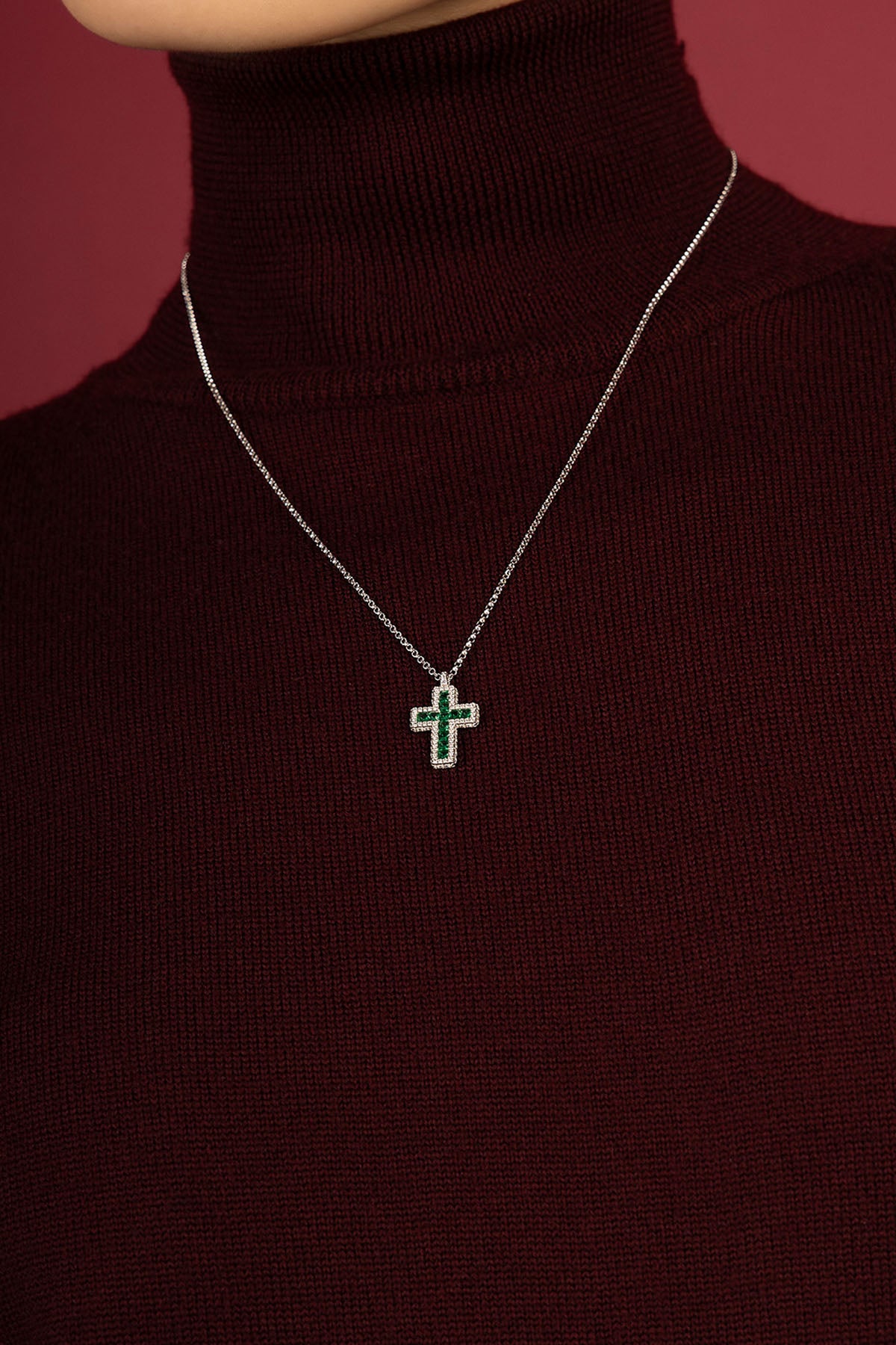 Woman wearing APM cross necklace, a quiet emerald glow against burgundy.