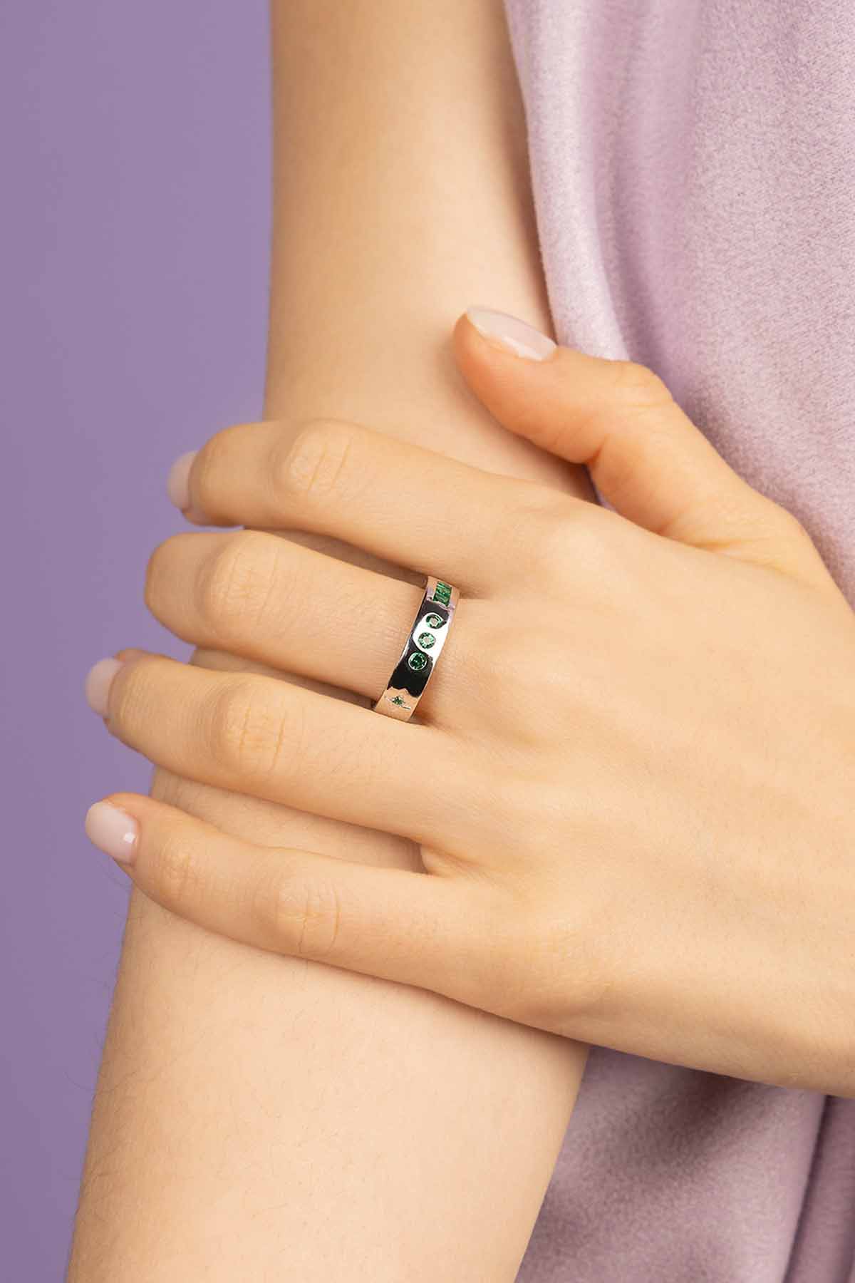 Close-up of a hand wearing a ring with green gemstones on a purple background