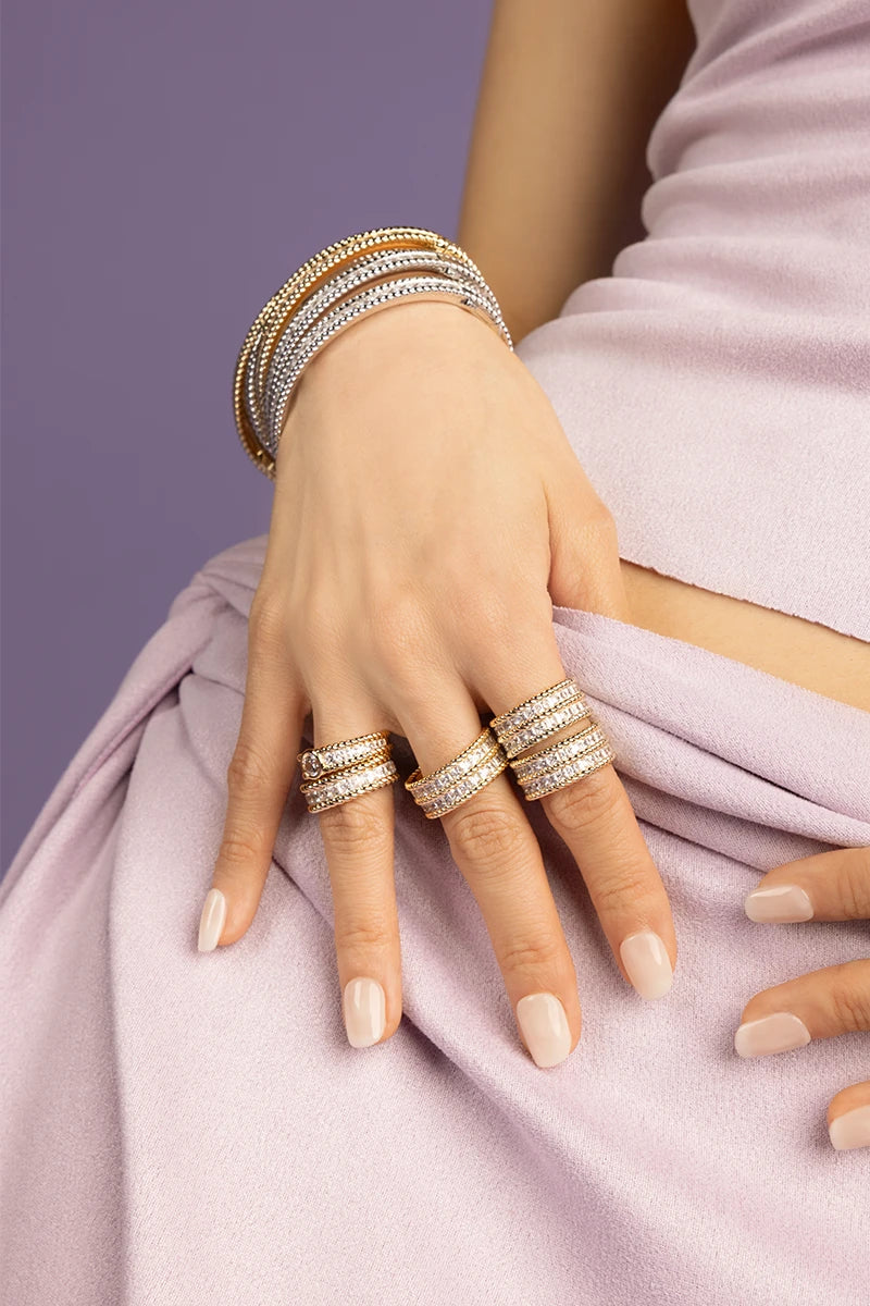 Hand wearing multiple yellow gold and silver rings from APM MONACO Printemps collection on a light purple fabric background