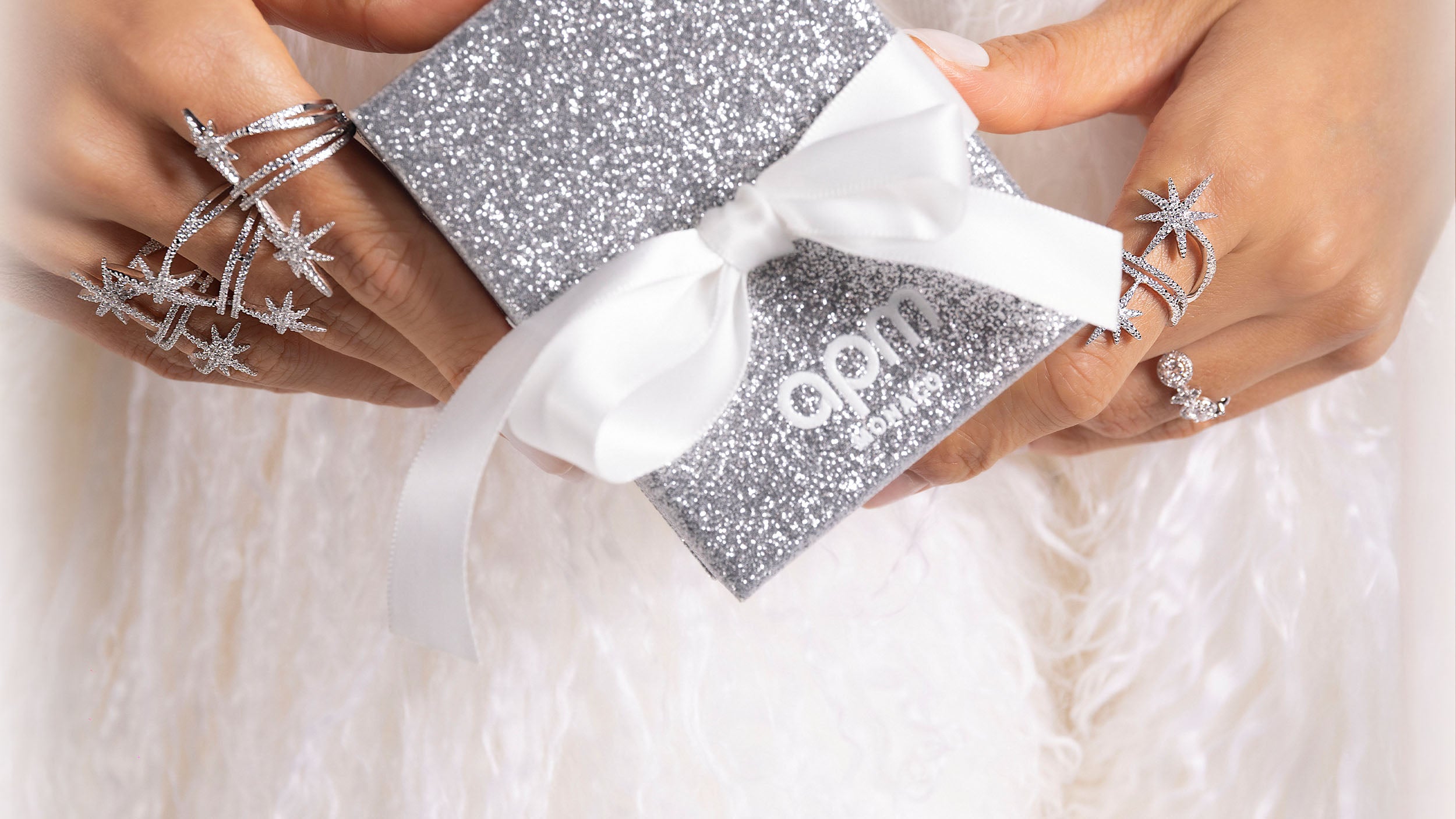 APM MONACO silver glittery gift box with a white ribbon held by hands wearing star-shaped rings on a light background.