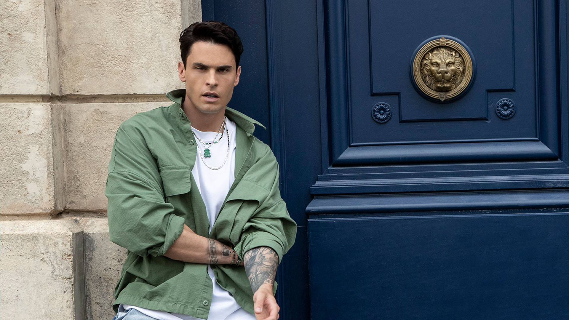 Man in green jacket and with APM MONACO yummy collection necklace leaning against a blue door 